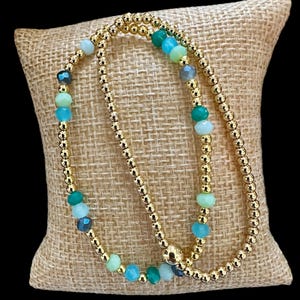 May include: Two gold beaded bracelets with green, blue, and clear gemstone beads.