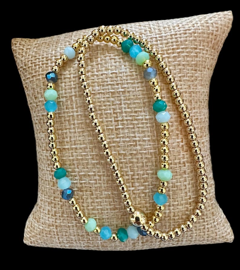 May include: Two gold beaded bracelets with green, blue, and clear gemstone beads.
