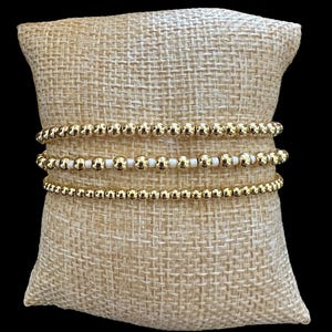 Puede incluir: Tres pulseras de cuentas doradas expuestas sobre un cojín de arpillera beige. Dos pulseras están hechas de cuentas de oro macizo, y la tercera pulsera tiene una mezcla de cuentas doradas y blancas. Las pulseras están apiladas.