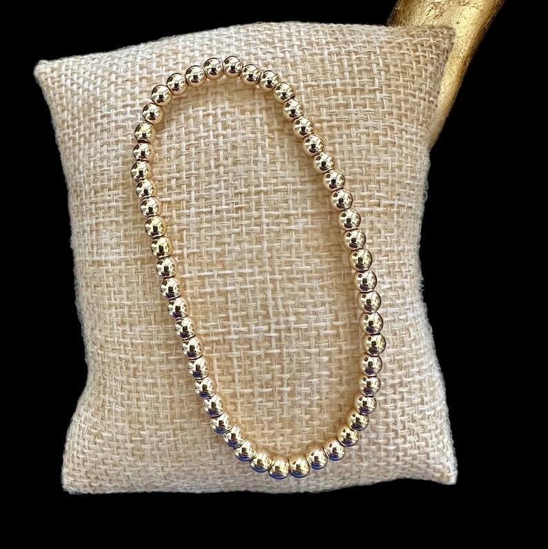 May include: A gold beaded bracelet on a beige fabric background.