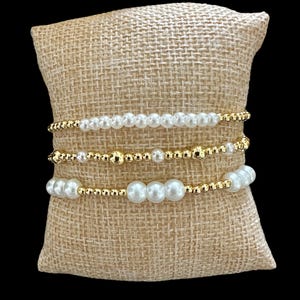 Può includere: Un set di quattro braccialetti con una combinazione di perle bianche e perline dorate. I braccialetti sono impilati su un cuscino di juta.