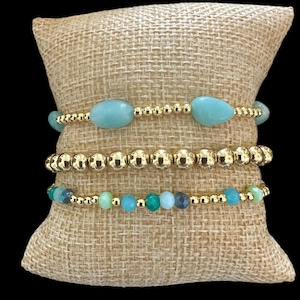 May include: Four gold bracelets with turquoise and green gemstone beads on a burlap background.