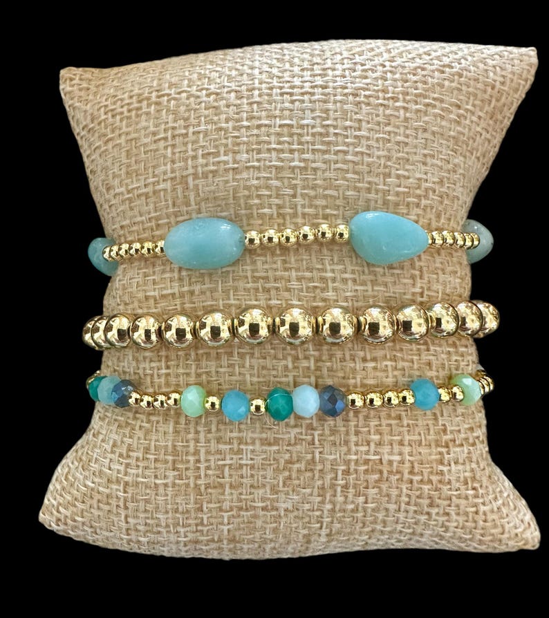 May include: Four gold bracelets with turquoise and green gemstone beads on a burlap background.
