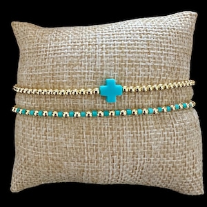 May include: Two beaded bracelets. The top bracelet is gold-colored with a turquoise cross charm. The bottom bracelet is turquoise with gold-colored beads. The bracelets are displayed on a burlap pillow.