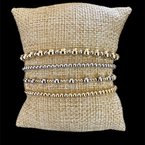 May include: A set of five bracelets with alternating gold and silver beads on a burlap background.