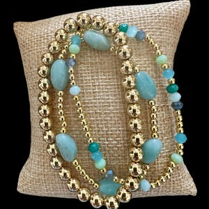 May include: Three gold beaded bracelets with green and blue gemstone beads. The bracelets are stacked on a burlap surface.