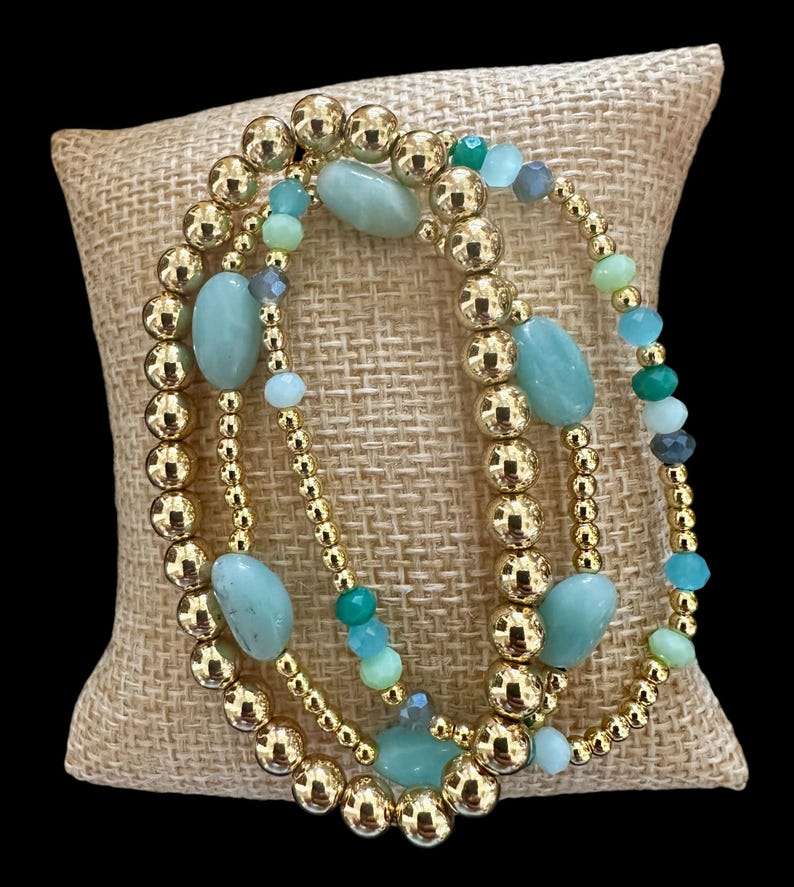 May include: Three gold beaded bracelets with green and blue gemstone beads. The bracelets are stacked on a burlap surface.