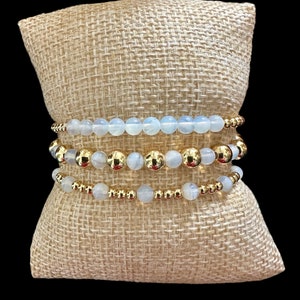 Pode incluir: Um conjunto de quatro pulseiras com contas de pedra da lua branca e detalhes dourados. As pulseiras estão empilhadas em um almofada de juta.