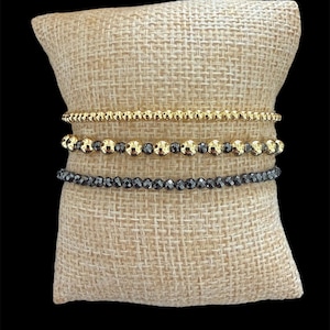 Puede incluir: Tres pulseras de cuentas doradas y gris oscuro se exhiben sobre una almohada de arpillera beige. La pulsera superior es dorada con pequeñas cuentas redondas. La pulsera del medio tiene cuentas doradas y gris oscuro. La pulsera inferior es gris oscuro con cuentas facetadas.