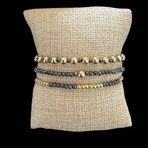 May include: Three beaded bracelets on a burlap cushion. The bracelets have gold and black beads. The top and bottom bracelets have gold beads, while the middle bracelet has black beads. The bracelets are stacked together.
