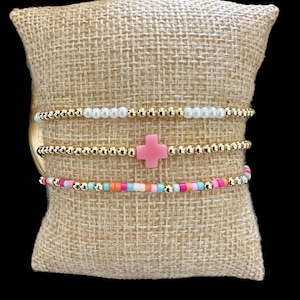 Puede incluir: Un conjunto de tres pulseras con cuentas doradas, perlas blancas y cuentas de colores. La pulsera del medio tiene un dije de cruz rosa.