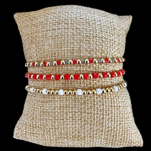 May include: Three beaded bracelets in gold, red, white, and gray. The bracelets are displayed on a beige burlap pillow against a black background. The beads are small and round, with gold-colored beads interspersed.