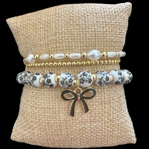 Puede incluir: Tres pulseras doradas con un patrón floral blanco y negro y un dije de lazo dorado. Las pulseras están apiladas sobre un cojín de arpillera.
