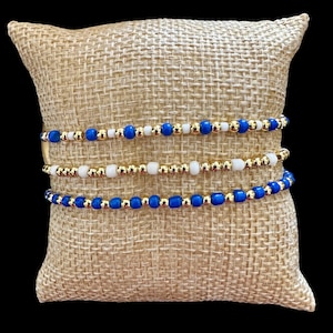 May include: Three gold beaded bracelets with blue and white beads. The bracelets are stacked on top of each other on a brown burlap surface.