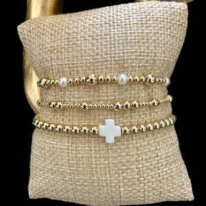 May include: Three gold beaded bracelets with a white cross charm. The bracelets are stacked on a burlap cushion.
