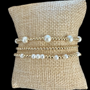 May include: Three gold beaded bracelets with white pearl accents are displayed on a beige burlap pillow. The bracelets vary in design, with some featuring larger pearls and others smaller ones. The gold beads add a touch of elegance.