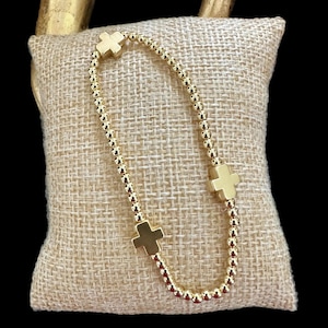 May include: A gold beaded bracelet with three gold cross charms. The bracelet is on a beige fabric background.