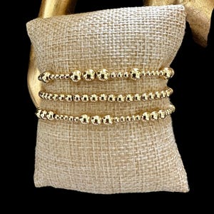 May include: A set of three gold beaded bracelets stacked on a beige fabric surface. The bracelets are made of small, round beads that are evenly spaced.