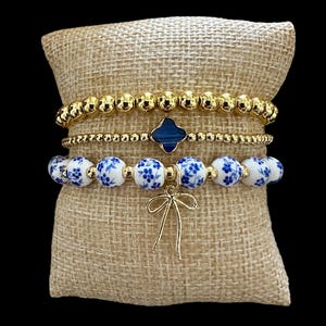 Puede incluir: Cuatro pulseras exhibidas sobre un cojín de arpillera. La pulsera superior es dorada con cuentas, seguida de una pulsera dorada con cuentas con un dije de trébol azul oscuro. La tercera pulsera tiene cuentas blancas con diseños florales azules y un dije de lazo dorado.