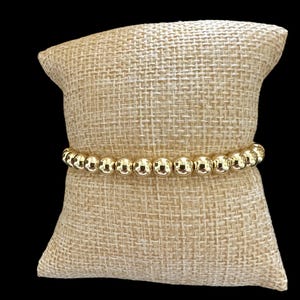 May include: A gold beaded bracelet with round beads on a beige fabric background.