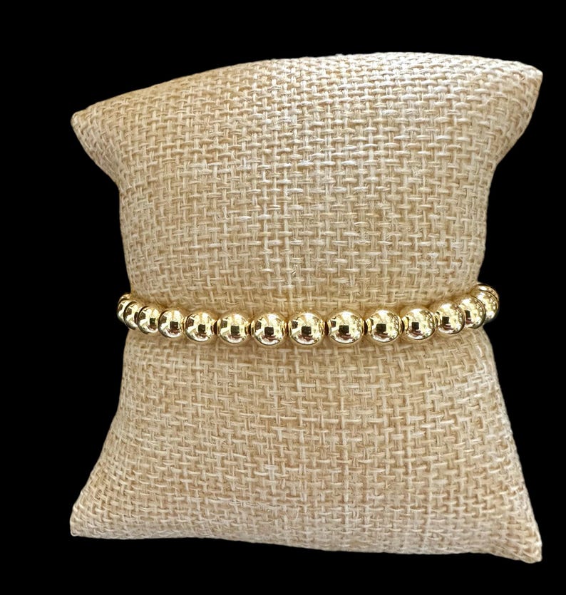 May include: A gold beaded bracelet with round beads on a beige fabric background.