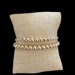 May include: Two gold beaded bracelets rest on a beige burlap pillow against a black background. The bracelets are made of small, round, shiny gold beads. The pillow has a woven texture.