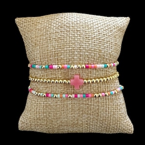 May include: A set of three stretch bracelets featuring small colorful beads in shades of pink, blue, and white, interspersed with metallic gold beads. The center bracelet includes a prominent pink cross-shaped charm as a focal point.