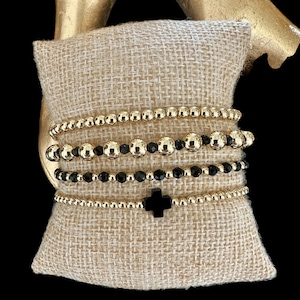 May include: A set of five bracelets featuring black and gold beads. The bracelets are stacked on top of each other and include a black cross charm.