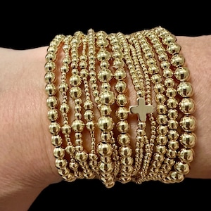 May include: A stack of gold beaded bracelets with a small gold cross charm. The bracelets are worn on a person's wrist.