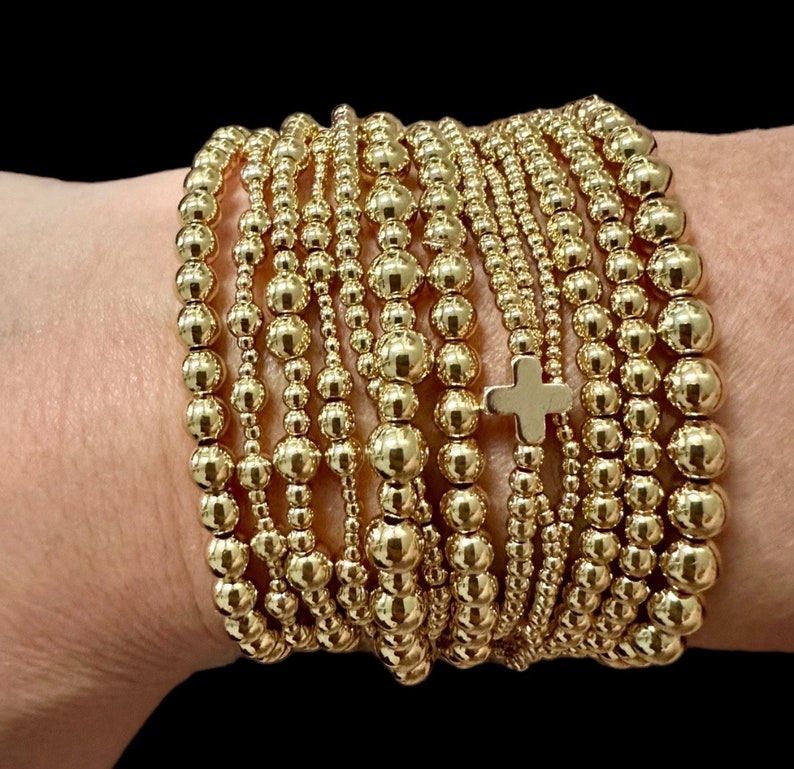 May include: A stack of gold beaded bracelets with a small gold cross charm. The bracelets are worn on a person's wrist.