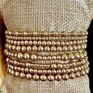 May include: A stack of gold beaded bracelets on a beige fabric surface.