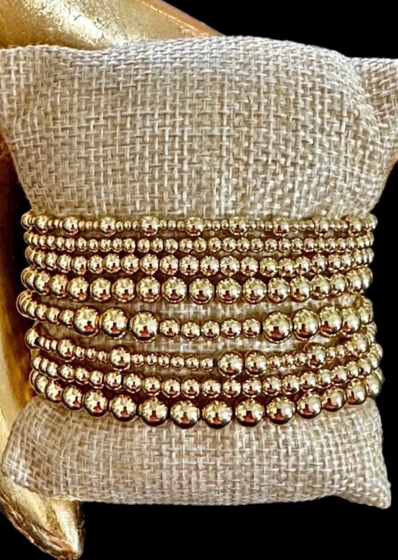 May include: A stack of gold beaded bracelets on a beige fabric surface.