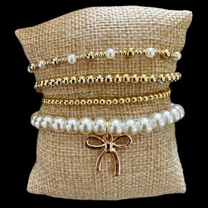 Puede incluir: Un conjunto de cuatro pulseras de cuentas doradas y blancas con un dije de lazo dorado. Las pulseras se muestran en un cojín de arpillera.