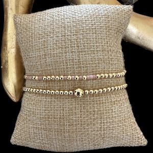 以下が含まれることがあります： ピンクのビーズのブレスレットを重ねた、2つの金のビーズのブレスレット。ブレスレットは、茶色の麻のクッションの上に置かれています。