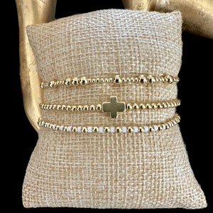 May include: Four gold beaded bracelets stacked on a beige burlap cushion. The top bracelet features a gold cross charm.