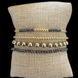 May include: Four beaded bracelets on a burlap pillow. The bracelets alternate between gold and dark grey beads. The beads are round and faceted, creating a sparkling effect. The bracelets are stacked together.