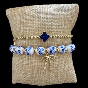 Puede incluir: Un conjunto de dos pulseras con cuentas de flores azules y blancas y una cadena dorada con un dije de trébol azul. Las pulseras están sobre una superficie de tela beige y una de las pulseras tiene un dije de lazo dorado.