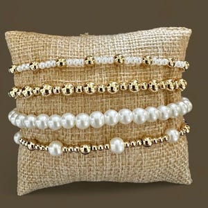 May include: Five bracelets stacked on a burlap cushion. The bracelets are made of gold beads and white pearls.