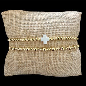 May include: Two gold beaded bracelets with a white cross charm are displayed on a tan burlap pillow. The bracelets are made of small, round, gold-colored beads. The white cross charm is centered on the top bracelet.