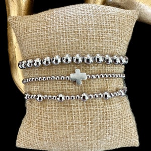 May include: A set of three silver beaded bracelets with a silver cross charm. The bracelets are stacked on a brown burlap cushion.