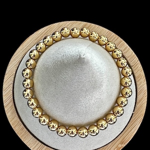 May include: A gold beaded bracelet with a shiny finish. The bracelet is displayed on a wooden stand.