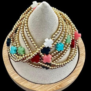 May include: A stack of gold bead bracelets with colorful cross charms. The bracelets are displayed on a wooden stand.