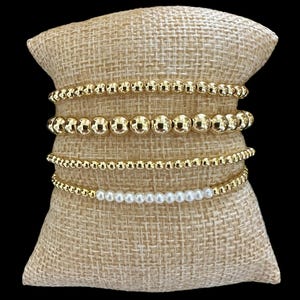 May include: A set of five gold bead bracelets stacked on a burlap surface. The top bracelet is a single strand of gold beads, the second bracelet is a double strand of gold beads, the third bracelet is a single strand of gold beads, the fourth bracelet is a single strand of gold beads, and the bottom bracelet is a single strand of white pearl beads.