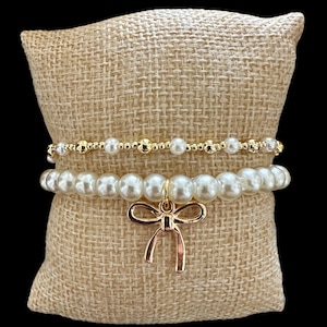 Puede incluir: Dos pulseras de perlas con un dije de lazo dorado. Las pulseras están sobre un fondo de arpillera marrón.