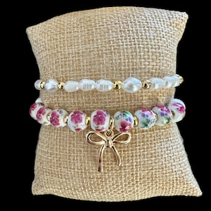 Puede incluir: Tres pulseras apiladas sobre un cojín de arpillera. La pulsera superior es una hilera de perlas blancas con detalles dorados. La pulsera del medio es una hilera de cuentas blancas con diseños florales rosas. La pulsera inferior es una hilera de cuentas blancas con diseños florales rosas y un dije de lazo dorado.
