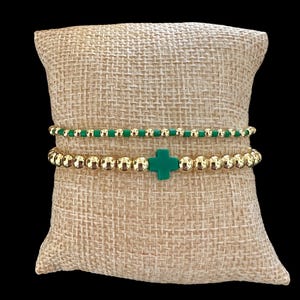 May include: Two beaded bracelets. The top bracelet has small green beads. The bottom bracelet has gold-colored beads and a green cross charm. The bracelets are displayed on a tan, textured pillow against a black background.