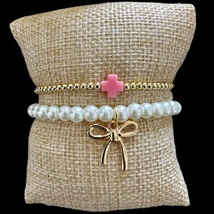 Puede incluir: Dos pulseras de oro con un dije de cruz rosa y un dije de lazo dorado. Las pulseras están sobre un fondo de arpillera marrón.