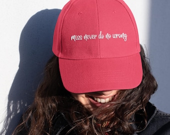 May include: A red baseball cap with the phrase "miss never do no wrong" in white script. The cap is made of a soft fabric and has a curved brim. The person wearing the cap is smiling.