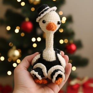 May include: A handmade crochet goose wearing a black and white hat and overalls. The goose has an orange beak and feet, and black eyes. The background features a Christmas tree with red and gold ornaments.