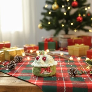 May include: A handmade crochet cupcake-shaped container with a green base and a cream-colored top, adorned with red cherry-like embellishments. A red and white checkered bow is tied on the front. The container sits on a red and green plaid tablecloth, with a Christmas tree and wrapped gifts in the background.